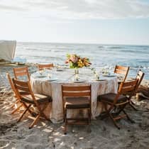 A wedding reception table adorned with glassware and a floral centerpiece is set up on a sandy beach by the ocean.