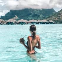 A person standing in clear turquoise water, holding a coconut drink, with overwater bungalows and mountains in the background