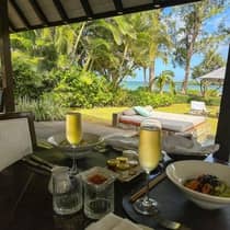 An outdoor table with food and two chilled glasses of champagne overlooking a pool, daybed and ocean view