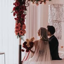 A couple in wedding attire with a bouquet of white and red roses, sitting on a bed adorned with red and pink flowers and a silver glitter chandelier decoration