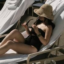 A woman in a black swimsuit and crochet sunhat sits on a wicker lounge chair covered with a white pool towel
