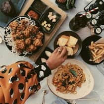 Table spread with various dishes, including fried chicken, noodles, bread rolls, fries and dipping sauces, with a person reaching for food