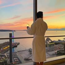 A guest dressed in a plush white robe and holding a glass of white wine stares out a window overlooking the Seattle sunset on the water.