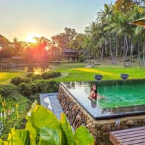 Person relaxing in an infinity pool overlooking rice fields and a pond at sunset, surrounded by lush tropical greenery.