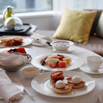 An In-Room Dining breakfast featuring eggs Benedict, fresh fruit, yoghurt and coffee, elegantly arranged on a table by a window with soft pillows in the background.