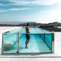 Man stands in raised glass swimming pool on white patio under blue sky