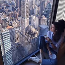Woman with coffee cup sits in large window overlooking New York City rooftops