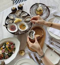 Top view of a dining table with a seafood platter featuring oysters on ice, dipping sauces, salad and bread, as a person prepares to eat an oyster.