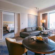 A hotel suite featuring a cozy living area with a sofa, a dining table with fresh fruit and a separate bedroom visible through sliding doors, lit by a chandelier.