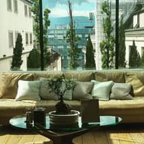 Beige sofa with decorative pillows, a small bonsai plant on a glass coffee table and a glass wall overlooking city buildings, trees and distant mountains
