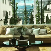 Beige sofa with decorative pillows, a small bonsai plant on a glass coffee table and a glass wall overlooking city buildings, trees and distant mountains