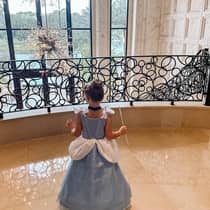 A young girl poses in a princess costume on a balcony overlooking a wall of glass windows
