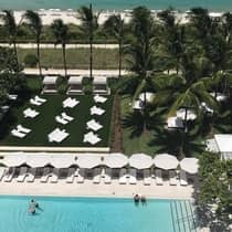 Aerial view of a beachfront pool area with white lounge chairs, umbrellas, cabanas, palm trees, and the ocean in the background.
