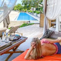 Woman wearing swimsuit lounges on patio cabana bed under white curtains near swimming pool