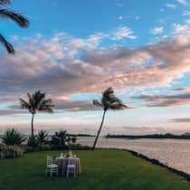 A private dining table for two at sunset, located oceanside on a green patch of grass, surrounded by palm trees and greenery