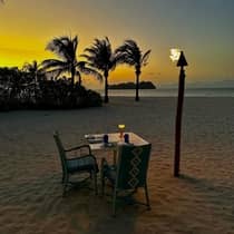 A dining table set for two on the beach at sunset, lit by a torch and candlelight