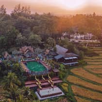 Aerial view of a resort with terraced pools, sun loungers and traditional architecture surrounded by rice fields at sunset.