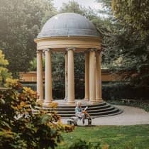 Circular pavilion with columns and a domed roof in a garden, surrounded by trees, with a person seated on a bench reading.