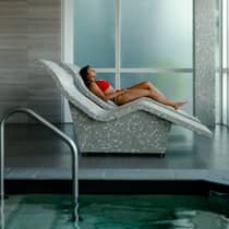A woman dressed in a red swimsuit lays on a mosaic tile lounge chair next to a spa pool