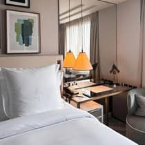 The corner of a room with a plush bed, framed artwork above and a glass nightstand with a modern pendant light hanging above