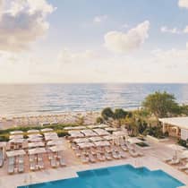 Aerial view of an outdoor swimming pool, rows of chairs, and umbrellas on the deck near the beach and ocean, creating a picturesque seaside escape.