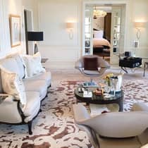 Luxury hotel suite living area with a sofa, modern chairs, round coffee table, desk, patterned rug and doors leading to a bedroom with a king bed