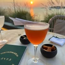 A frothy, orange cocktail topped with a mini decorative paper airplane, served by a sea view at Avra Bar