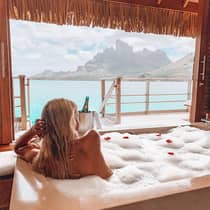 A person relaxing in a bubble bath with an ocean and mountain view from a deck, with a bottle of champagne nearby