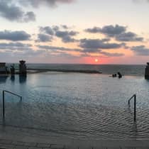 Silhouettes of three people swimming at edge of infinity pool, ocean and sunset in background