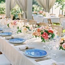 Close up of a wedding reception table lined with colorful floral arrangements, blue dining plates and white linens.