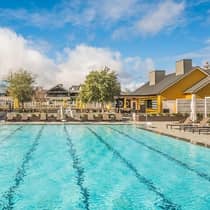 Outdoor swimming pool with lounge chairs and umbrellas, surrounded by buildings and trees under a partly cloudy sky.