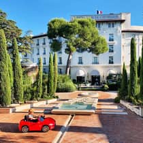 Young child rides in small red toy car in hotel courtyard with fountains, landscaped trees