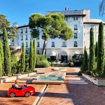Young child rides in small red toy car in hotel courtyard with fountains, landscaped trees