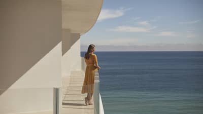 A woman standing on a balcony of a white stone building with the ocean in the background.