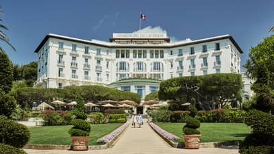 Two people walk down sunny stone path under large Grand-Hotel du Cap-Ferrat