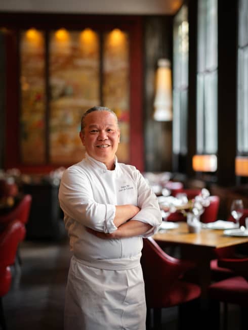 A male chef in a chef's coat in a restaurant with red chairs.