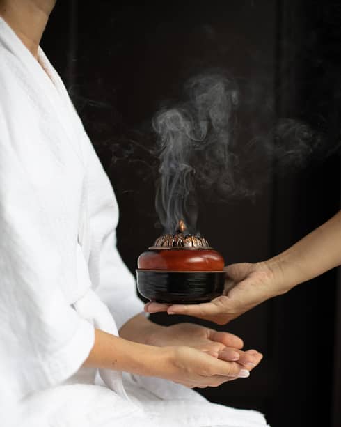 A person in a white robe receiving a steaming incense bowl from a therapist, creating a calming ambiance during a spa treatment.