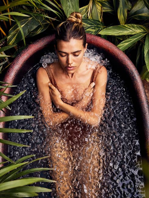 A guest, wearing a bathing suit, is sitting and relaxing in a rustic bathtub full of ice with foliage encircling the tub.