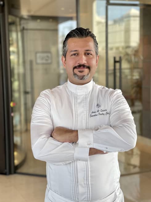 A man stands in front of a glass door in a white Four Seasons chef uniform.