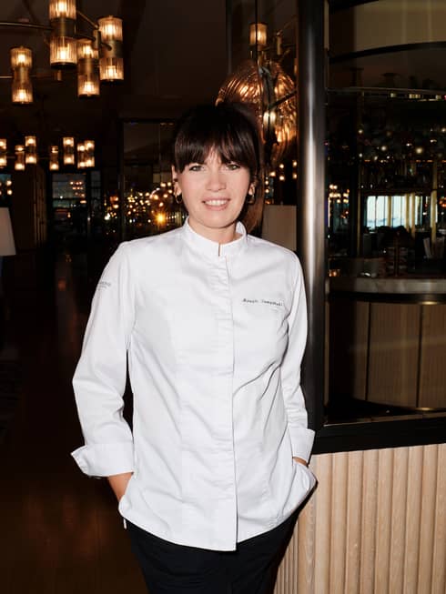 Chef Maggie Tampakaki, standing in white chef?s coat with hands in pockets