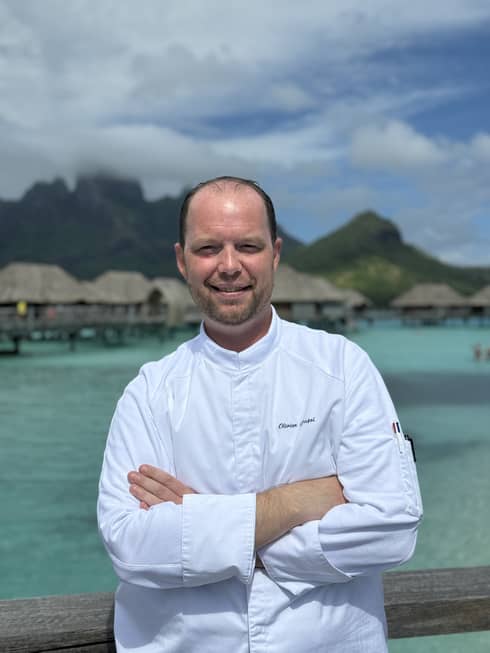Smiling chef in white coat stands with arms crossed against backdrop of overwater pavilions