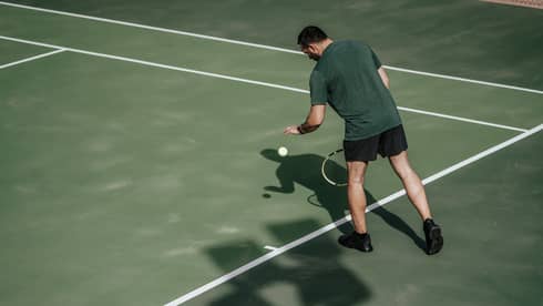 Man on tennis court bouncing tennis ball with left hand, preparing to serve