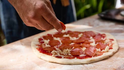 Close-up of a hand placing pepperoni on a small, round, handmade pizza that is placed on a floured wooden surface.