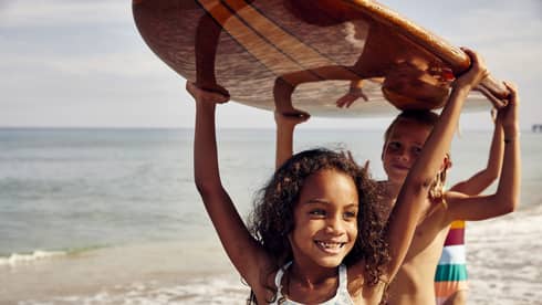 Three young children carry a surfboard on the beach, smiling and enjoying the sunny day. The ocean is in the background.