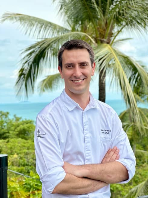 Person in white chef's coat with Four Seasons logo stands outdoors with arms crossed, palm tree in background
