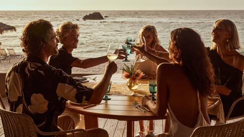 Five guests raise their glasses in a toast around a round wood table on a wood deck overlooking the beach and ocean.