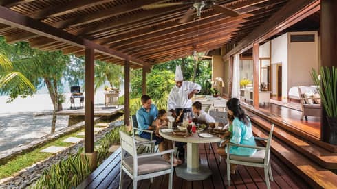 Chef in uniform presents meat skewer to family around table on Beach Villa patio