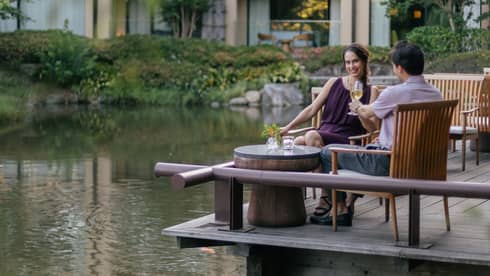 Smiling couple clink champagne flutes at a café table on a wood deck overlooking a rippling pond bordered by lush foliage.