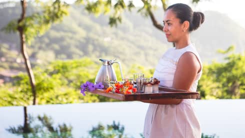 Woman carries breakfast tray with tropical flowers past outdoor pool