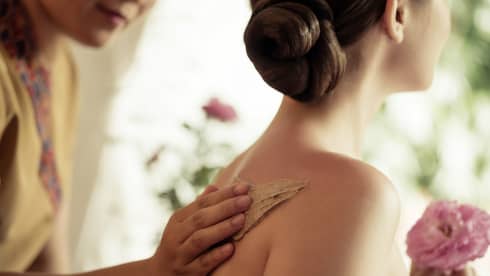 Spa staff rubs thick lotion on woman's bare shoulders as she holds a fresh flower